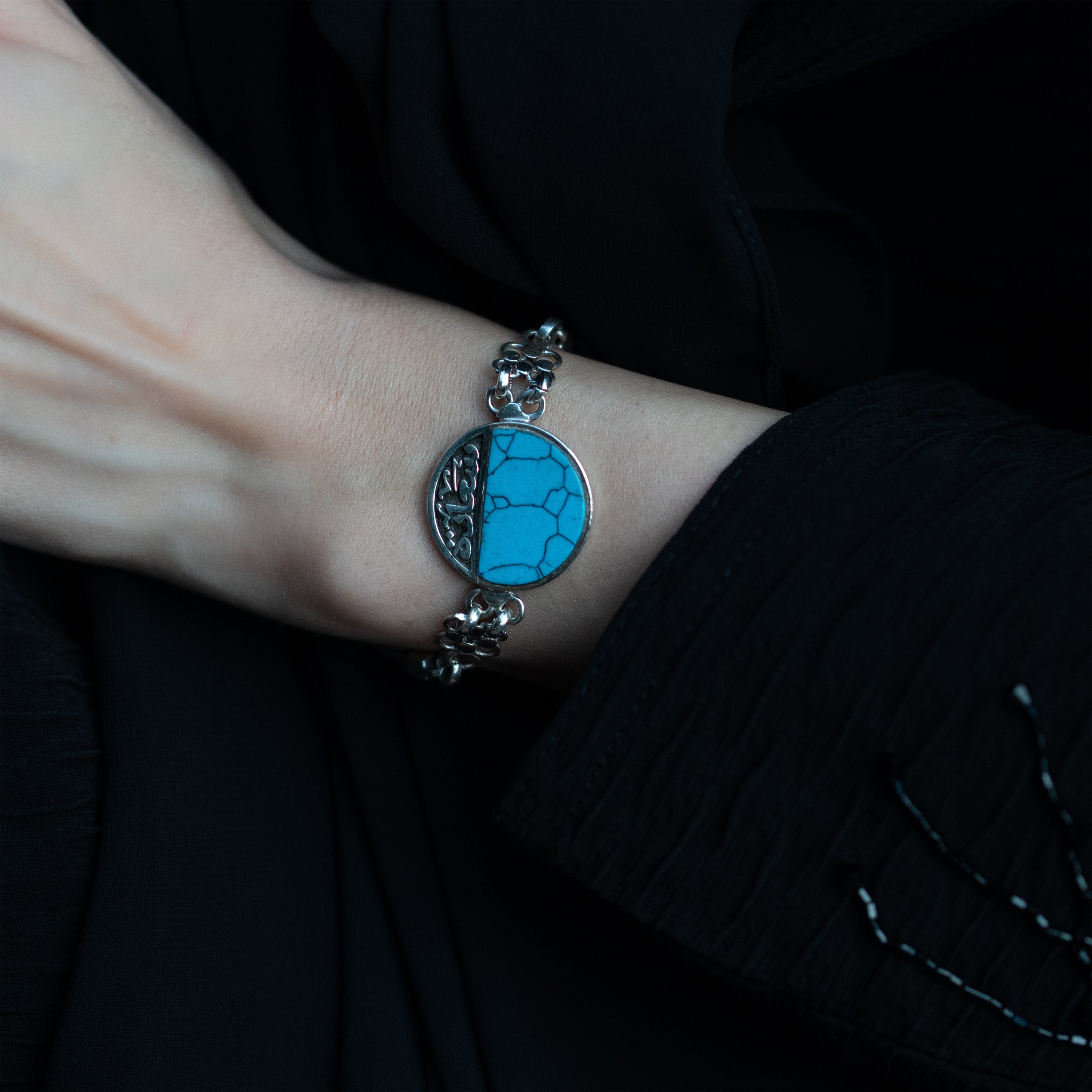 A person's wrist wearing a silver bracelet with a turquoise stone in the center, surrounded by two silver chains on each side with a toggle clasp at the end.