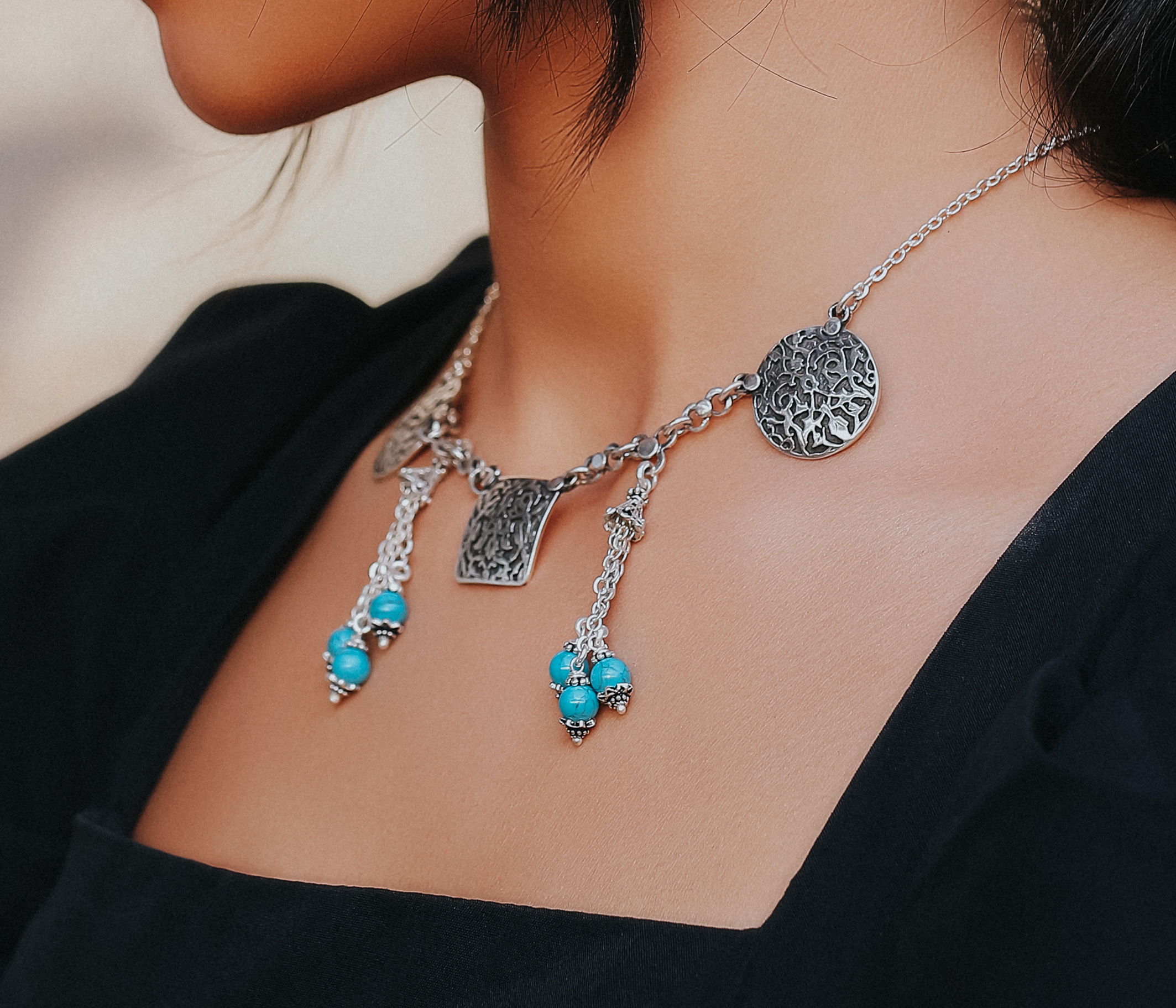 A close-up image of a silver necklace with oxidized details, featuring an engraved square shape in the middle, chain links, and three turquoise stones on the sides, with an engraved circle also on both sides.